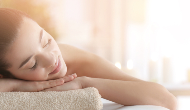 Eine Frau liegt in Bauchlage auf eine Massageliege. Ihre Hände unter dem Kopf, stützende auf einem Handtuch neigt Sie Ihrem Kopf leicht auf die Seite und lächelt ganz entspannt. Im Hintergrund schein die Sonne und vermittelt ein Gefühl der Geborgenheit.