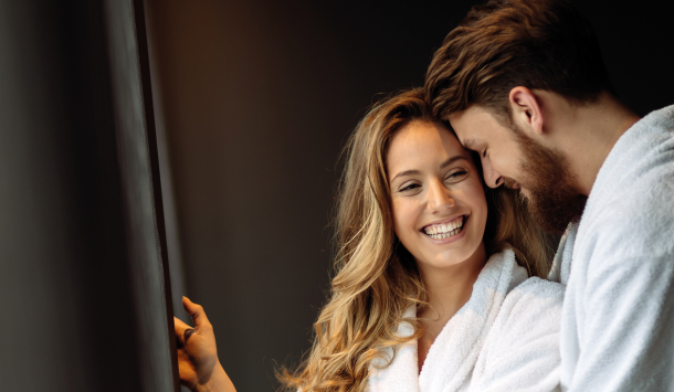 Pärchen in Bademäntel kuscheln im stehen an einander. Sie lächeln sich gegenseitig an.