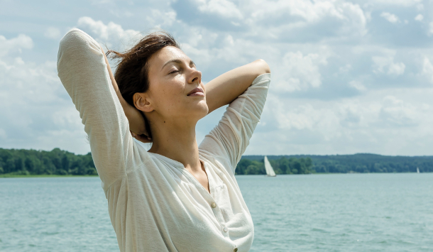 Frau steht am Wasser mit Ihren Händen gestreckt hinter dem Kopf. Im Hintergrund sieht man den See mit einem Segelboot auf dem Wasser.