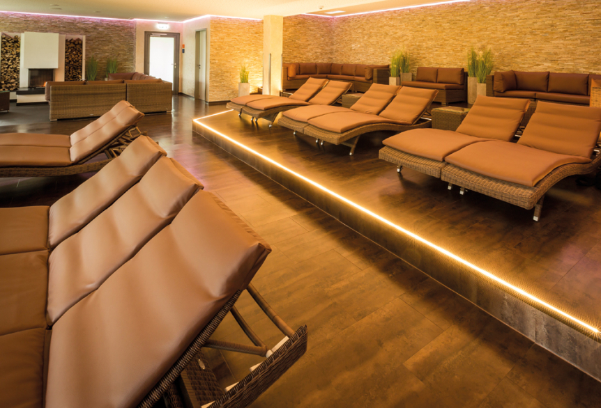 Ruheraum in der PanoramaSauna mit Liegen auf denen sich braune Auflagen befinden. Alle Liegen sind in die selbe Richtung ausgerichtet. Im Hintergrund auf dem Podest befinden sich Sitzecken mit Kissen versehen und als dekorative Elemente befinden sich grüne Dekopflanzen. Eine Kaminecke mit Feuer ist im Hintergrund zu sehen. Der Raum wird mit warme weißen Deckenspots und einem integrierten Lichtschlauch im Podest beleuchtet.