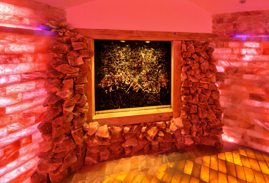 Raum mit warmem, rötlich-orangem Licht, dessen Wände aus Salzsteinen bestehen. In der Mitte befindet sich ein beleuchtetes Wandbild aus Kristallen oder Salzbrocken. Der Boden reflektiert das Licht und erzeugt eine gemütliche, entspannende Atmosphäre – typisch für eine Salzgrotte oder ein Salztherapie-Zimmer.