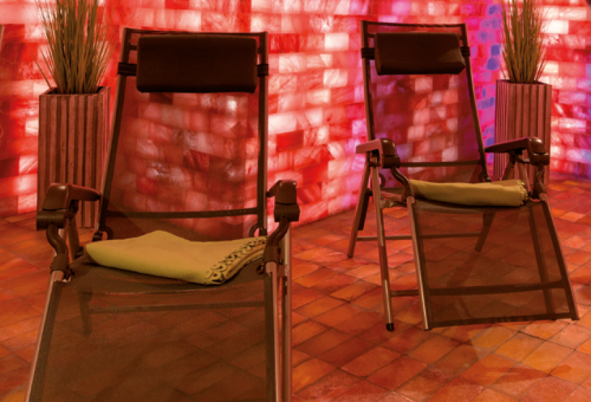 Mehrere Relaxliegen stehen in einem stimmungsvoll beleuchteten Ruheraum mit roten Salzwänden und warmem Licht. Der Boden ist mit Salzsteinen ausgelegt, an den Wänden stehen dekorative Pflanzen.