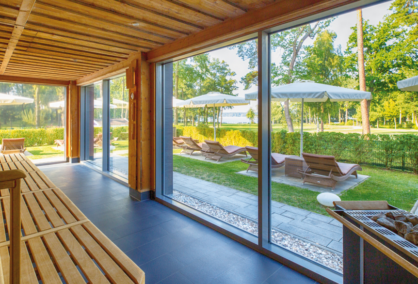 Abgebildet ist die KeloSauna der PanoramaSauna mit Blick auf den grünen Kurpark in Bad Saarow, mit Blick auf den blauen Scharmützelsee. Die Saunadecke und die Saunabank, sind mit groben Holzstrukturen versehen. Der Fußboden besteht aus dunkel grauen Fließen. Eine riesige Glasfront lässt es zu, dass der Raum Sonnendurchflutet wird und man sieht die braunen Außenliegen mit weißen aufgespannten Sonnenschirmen. Ein dunkel grauer Saunaaufgussofen ist am Rande des Bildes zu erkennen.