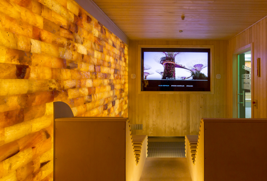 Ausschnitt aus der KinoSauna mit Blick auf den großen Bildschirm, auf dem ein Film abgespielt wird. Die Decke und die rechte Wand ist mit Holz verkleidet. Die linke Wand besteht aus Salzsteinen, die Gelb-Orangefarbendes Licht abgeben. Holzliegen dienen als Sitz- oder Liegemöglichkeit.
