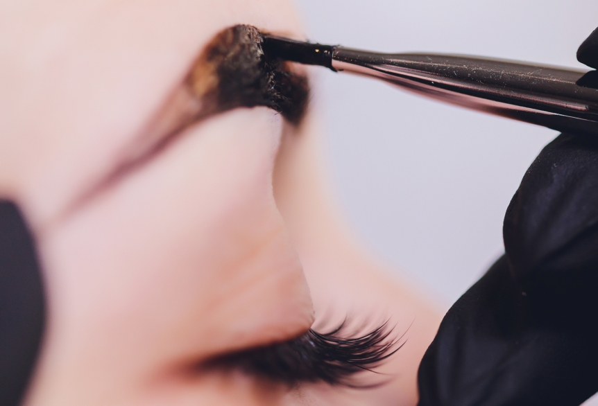 Nahaufnahme eines geschlossenen Auges einer Frau, die ihre Augenbrauen gefärbt bekommt. Mit einem schmalen Pinsel wird die dunkelbraune Farbe auf die rechte Augenbraue aufgetragen.  