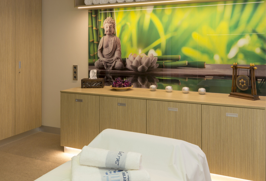 Ein hell erleuchteter Therapieraum mit Cremefarbenden Teppich. Im unteren Bereich des Bildes befindet sich eine Massageliege bedeckt mit einem weißen Laken und weißen Handtüchern mit der Aufschrift SaarowTherme. Im Hintergrund befindet sich eine helle Holzkommode mit Dekorativen Elementen wie einer Taschentücherbox, eine Schale mit Lila Blumen, 4 Teelichterständer und einem Chinesischen Gong Becken. Über der Kommode hängt ein hell grünes Bild mit einem Buddha, darüber ein Hängeschrank mit weißen Handtüchern