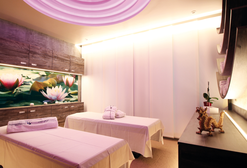 Ein Anwendungsraum mit zwei Massageliegen die mit weißen Laken bedeckt sind. Dekorativ befindet sich auf der einen Liege ein weißes Handtuch mit dem SaarowTherme Logo in Blau und auf der anderen ein weißer Bademantel mit Logo. Im Hintergrund sind graumelierte Hängeschränke mit einem Dekorativen Bild mit rosafarbenen Lotusblüten. Der Raum wird durch Deckenbeleuchtung und Unterschrankbeleuchtung warm weiß erleuchtet. Der Boden besteht aus einem Cremefarbenen Teppich. 