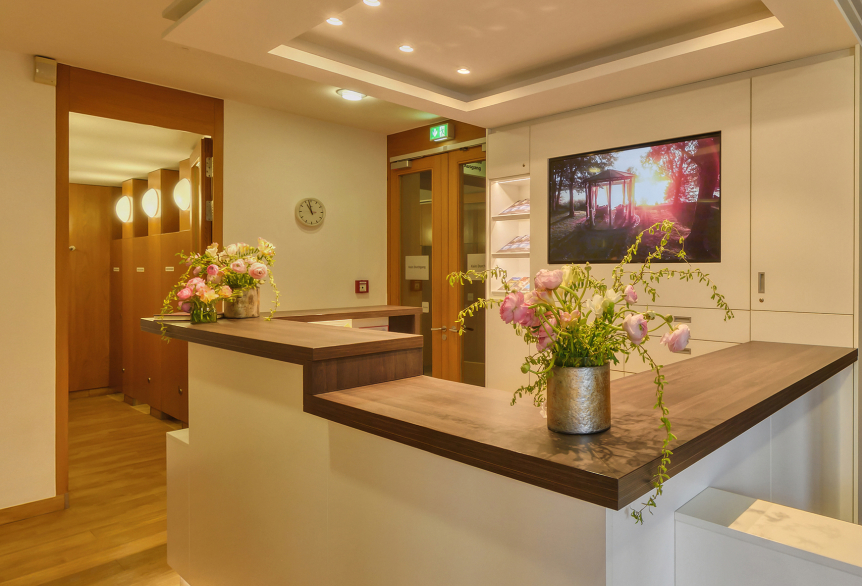 Terminierungsbereich im Wellnessbereich der SaarowTherme. Ein hoher weißer Tresen mit dunkelbrauner Holzplatte, dekorativ geschmückt mit Rosa Blumen und grünen hängenden Ästen. Im Hintergrund sieht man die Umkleidekabinen mit ihrer Holzoptik und einen Fernseher mit einem Bild am See und dem Sonnenuntergang. Der Tresen wird durch Deckenspots warm weiß beleuchtet. 