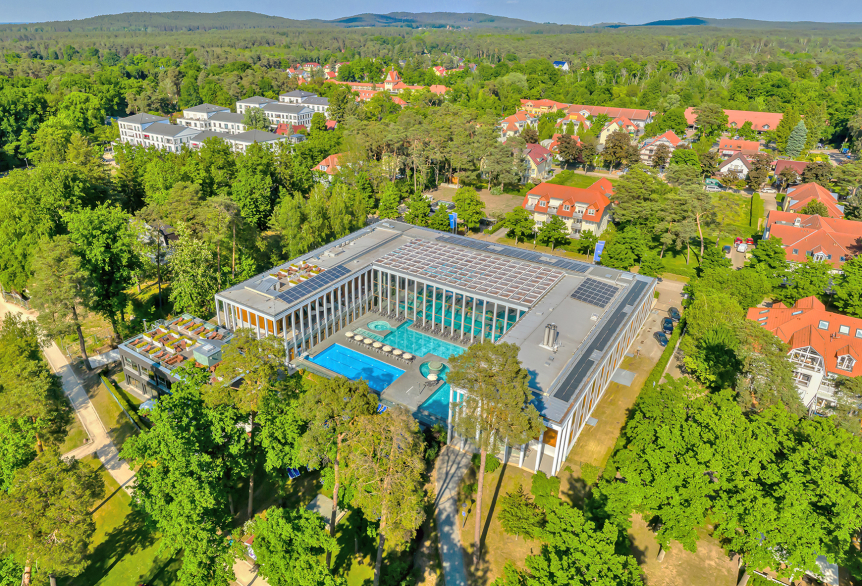 Das SaarowTherme Gebäude mit Blick auf die Außenpools aus der Vogelperspektive  und der grünen Umgebung des Kurparks, mit vereinzelten Wohnhäusern. 