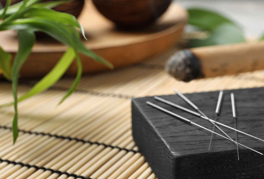 Ein Produktbild mit einer Bambusunterlage, auf der sich ein Auschnitt von einem schwarzen, schmalen Holzstück befindet auf dem silberfarbene Akupunkturstäbchen platziert wurden. Im Hintergrund befindet sich die Spitze eines angezündeten Moxstäbchen und dekorative Grünpflanzen mit einer Bambusschale.