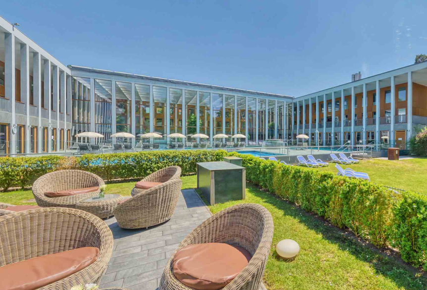 Außenbereich der PanoramaSauna mit Terrasse und Blick auf den Außenbereich des Thermalbades aus der Normalperspektive. Im Vordergrund befinden Sitzmöglichkeiten eingebettet von grünen Sträuchern. Im Hintergrund sieht man das Thermengebäude mit der großen Glasfront , mit weißen Säulen und Gebäudefront in Holzoptik. Aufgespannte weiße Sonnenschirme bei strahlendem blauen Himmel.