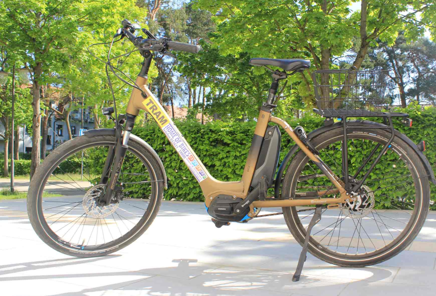 Fahrrad mit rotem Rahmen, schwarzem Lenker und Sitz, gelber Aufschrift HOLM, stehend im Ständer vor grüner Hecke und Bäumen.