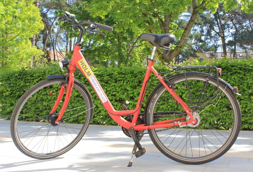 Fahrrad mit rotem Rahmen, schwarzem Lenker und Sitz, gelber Aufschrift ‚HOLM‘, stehend im Ständer vor grüner Hecke und Bäumen.