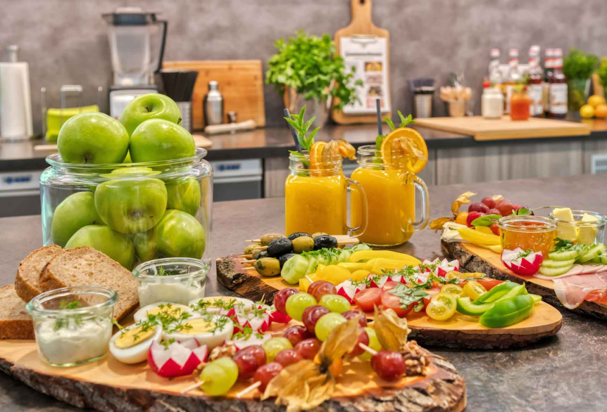 Auf einen Tresen angerichtete drei rustikale Brotbretter mit Speisen wie, Weintraubenspieße, gekochte halbierte Eier, Olivenspieße, Salami, Schinken und verschiedene Dips in kleinen Einweggläsern. Im Hintergrund sind dekorativ grüne Äpfel in einem großen Dekoglas und zwei Orangensäfte platziert.