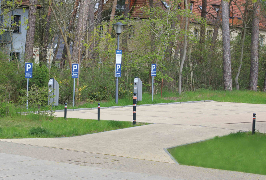 Zu sehen ist der E-Parkplatz an der SaarowTherme. Es sind vier blaue Schilder mit einem großen P ausgewiesen. Es sind vier graue Ladesäulen auf dem hellgepflasterten Parkplatz zu erkennen. Im Hintergrund befindet sich ein Wohngebiet hinter grünen Bäumen und Büschen versteckt. 