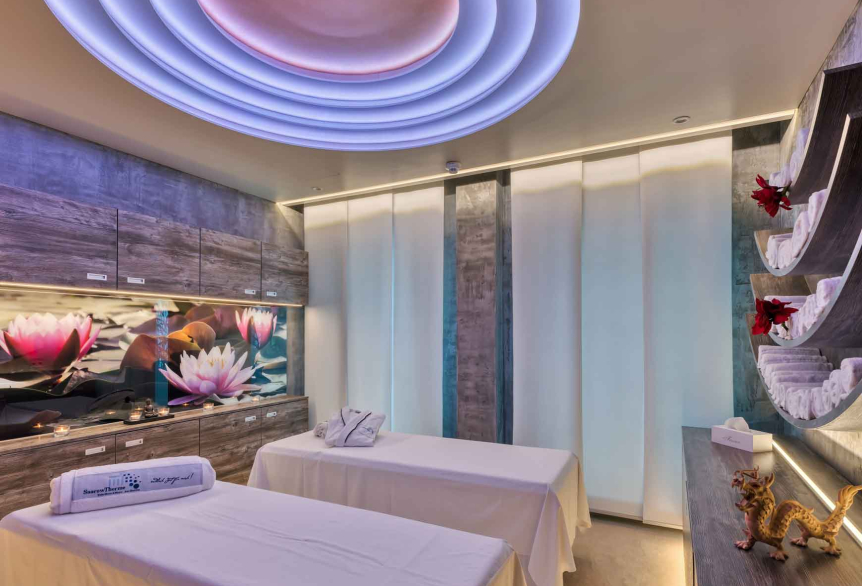 Ein Anwendungsraum mit zwei Massageliegen die mit weißen Laken bedeckt sind. Dekorativ befindet sich auf der einen Liege ein weißes Handtuch mit dem SaarowTherme Logo in Blau und auf der anderen ein weißer Bademantel mit Logo. Im Hintergrund sind graumelierte Hängeschränke mit einem Dekorativen Bild mit rosafarbenen Lotusblüten. Der Raum wird durch Deckenbeleuchtung und Unterschrankbeleuchtung warm weiß erleuchtet. Der Boden besteht aus einem Cremefarbenen Teppich. 