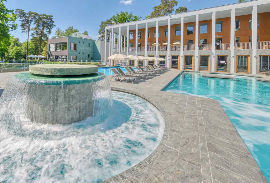 Außenbereich des Thermalsolebeckens  SaarowTherme am Tag mit plätschernden Strömungskanal, graue Liegen mit weißen aufgespannten Sonnenschirmen. Im Hintergrund ein Teil des Hauptgebäudes mit hellbrauner Gebäudefront und weißen Säulen im Vordergrund. Blauer Himmel mit weißen Schleierwolken. 