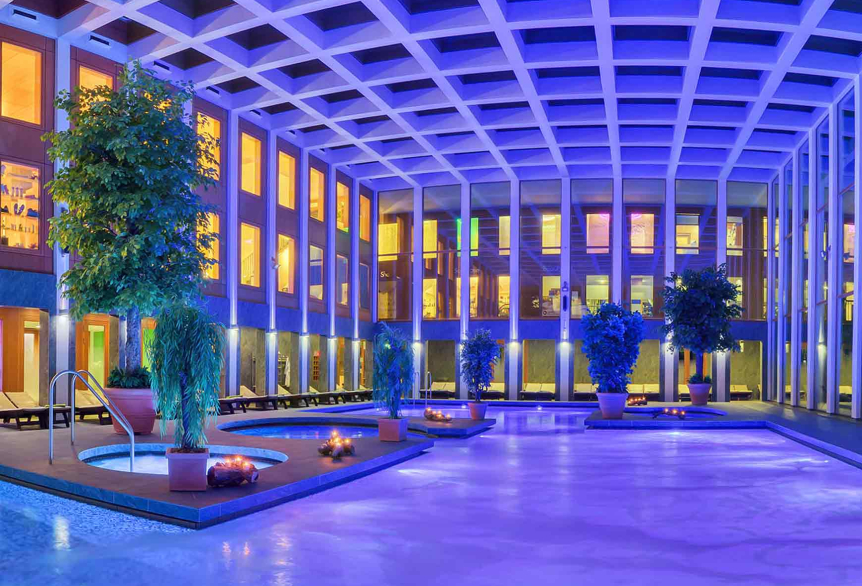 Abendlicher Innenbereich der SaarowTherme mit beleuchtetem Solebecken. Lila gefärbtes Wasser durch Unterwasserscheinwerfer, dekoriert mit Grünpflanzen und brennenden Kerzen am Beckenrand. Goldenes Licht fällt durch die Glasfront im oberen Bereich des Gebäudes.