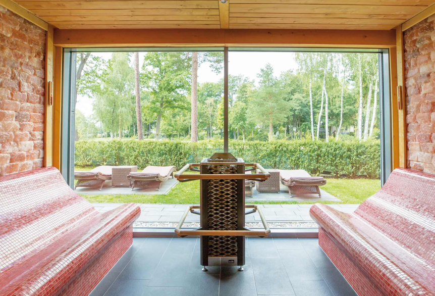 Aus  SalzSauna in der PanoramaSauna schaut man direkt auf den grünen Außenbereich mit braunen Liegen. Die Glasfront erhellt die Sauna mit natürlichem Sonnenlicht. Rechts und links von dem Metallfarbenen Saunaofen befinden sich jeweils rote, gewölbte Bänke aus kleinen Mosaikfliesen. Die Decke ist mit Holz abgedeckt und die Wände mit Salzsteinen versehen. 