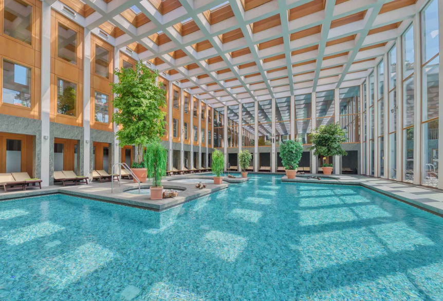 Thermalsolebad Innenbereich Tageslichtdurchflutet, türkisfarbenes Wasser. Dekorativ sind Grünpflanzen in verschiedenen Größen im Badbereich verteilt. Zahlreiche Liegemöglichkeiten aus dunklem Holz mit hellen Auflagen und grünen Decken. Eine große Glasfront durch die man den Außenbereich der SaarowTherme sehen kann.  