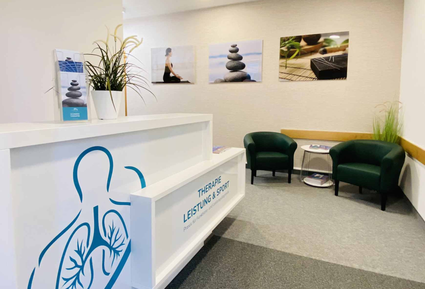 Zu sehen ist der Eingangsbereich der Arztpraxis an der SaarowTherme. Links vom Eingang befindet sich ein weißer Tresen mit Türkisfarbener Aufschrift "Therapie Leistung &Sport". Auf dem Tresen befindet eine kleine Grünpflanze und ein Prospektständer. Im Hintergrund befinden sich zwei dunkel grüne Sessel mit einem weißen, runden Couchtisch. Auf diesem befinden sich weitere Prospekte. Dekorativ ist der Eingangs Bereich mit einer Stehgrünpflanze und drei Bildern versehen. Hellgrauer Teppich, Cremefarbene Wand.