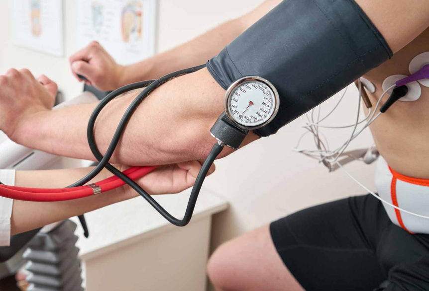 Ein Ausschnitt von einem Bild, wie ein Mann, Oberkörperfrei in einer schwarzen Sporthose auf einem Ergometer mit einem Blutdruckmessgerät am linken Oberarm und EKG Messgeräte am Oberkörper.