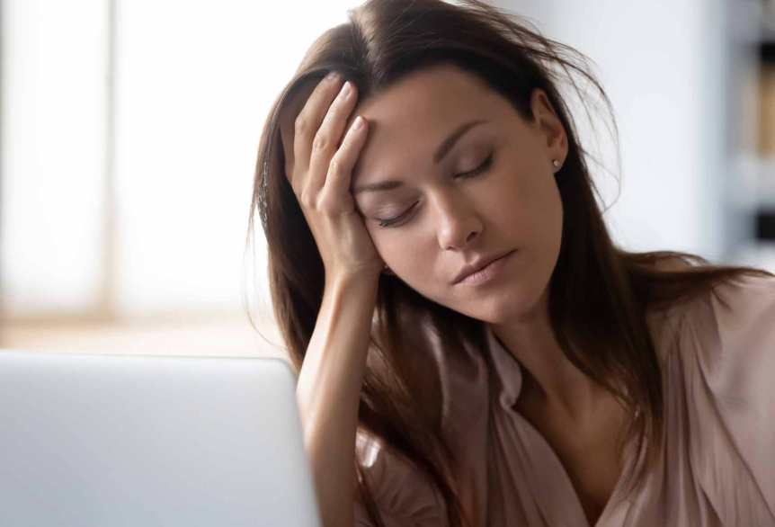 Eine Frau mit braunen, langen Haaren, sitzt an einem vollen Schreibtisch und stützt sich erschöpft den Kopf auf der Hand ab. Dabei hat sie ihre Augen geschlossen.