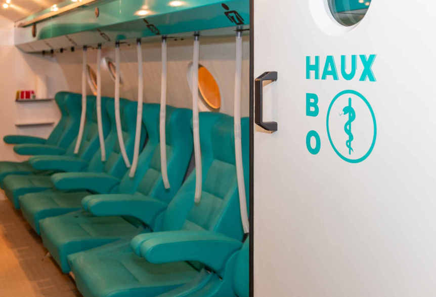 Ausschnitt der Druckkammer vom TL Tauchunternehmen Leunert. Aneinander gereihte, türkisfarbene Sitzmöglichkeiten, vergleichbar mit Sitzen in einem Zug, nur aus Leder. Weiße Schläuche für die Beatmung, hängen über den Sitzen. Eine weiße Tür mit der türkisfarbenen Aufschrift HAUX steht zu dem Innenraum der Druckkammer offen.