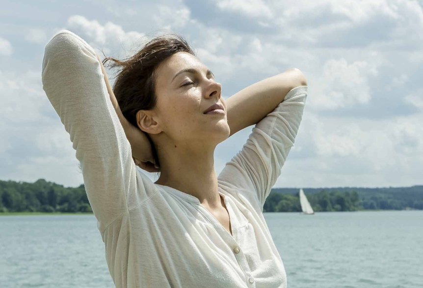 Eine Frau mit braunen Haaren und einem weißen langärmligen Shirt steht am See, verschränkt die Arme hinter dem Kopf und streckt ihr Gesicht in Richtung Sonne mit geschlossenen Augen. Im Hintergrund sind man Wald und davor den See mit Segelschiffen.