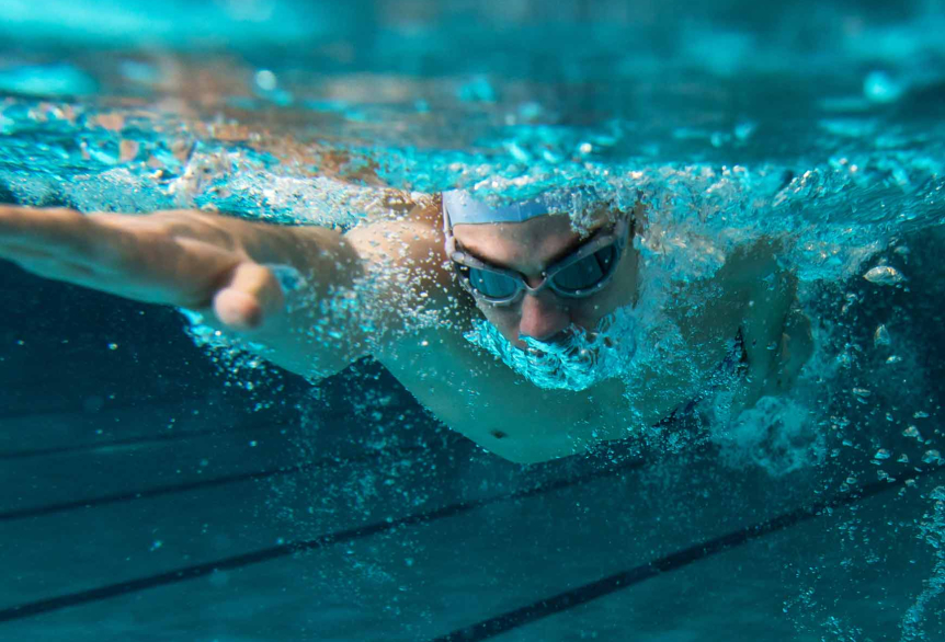 Ein Leistungsschwimmer mit Schwimmbrille und Badekappe wird unter Wasser in einem Sportbecken abgebildet. Er krault und hat seinen rechten Arm vorne. 