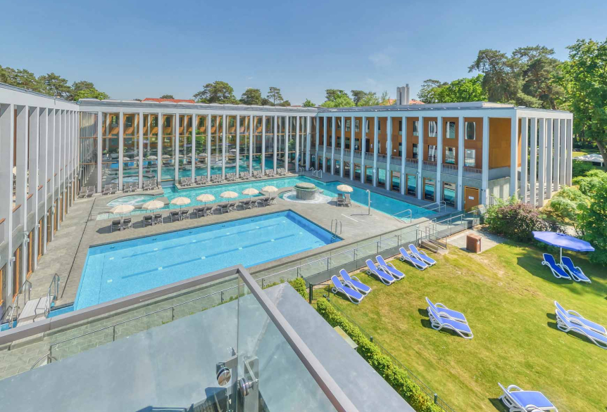 SaarowTherme Aussenbereich aus der Vogelperspektive mit grüner Liegewiese auf der blaue Liegen platziert sind und ein blauer Sonnenschirm. Sowohl das Türkisfarbene Sportbecken als auch das Solebecken im Außenbereich sind abgebildet. Drum herum ist das Thermalbadgebäude in umgekehrter U Form zu sehen mit weißen Säulen und hellbrauner Außenfassade. Am Außenbecken befinden sich Liegen mit weißen aufgespannten Sonnenschirmen.