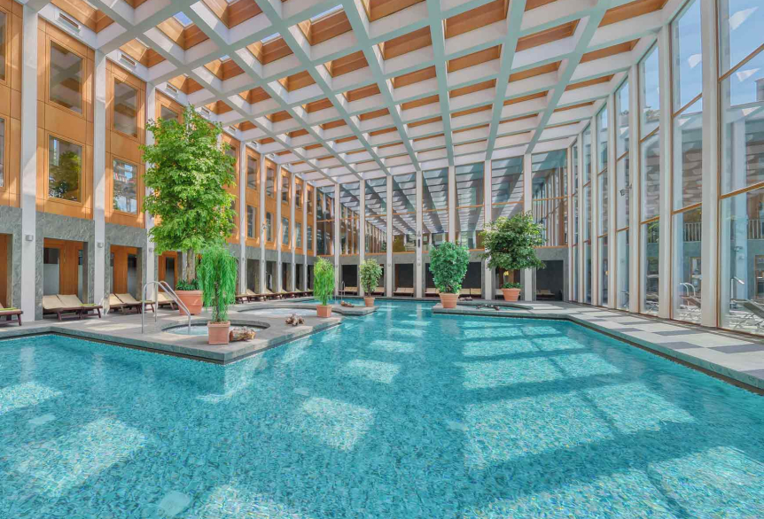 Thermalsolebad Innenbereich Tageslichtdurchflutet, türkisfarbenes Wasser. Dekorativ sind Grünpflanzen in verschiedenen Größen im Badbereich verteilt. Zahlreiche Liegemöglichkeiten mit grünen Decken und eine große Glasfront durch die man das Außenthermalsolebecken, Liegen und Sonnenschirme sieht. 