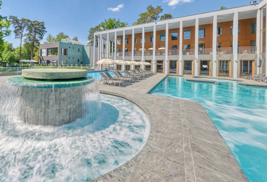 Außenbereich des Thermalsolebeckens  SaarowTherme am Tag mit plätschernden Strömungskanal, graue Liegen mit weißen aufgespannten Sonnenschirmen. Im Hintergrund ein Teil des Hauptgebäudes mit hellbrauner Gebäudefront und weißen Säulen im Vordergrund. Blauer Himmel mit weißen Schleierwolken. 