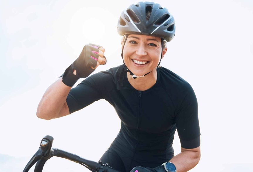 Eine Frau in kurzen, schwarzen Radsport Sachen  und einem schwarzen Helm auf einem Rennrad. Mit einer Hand am Lenker und die andere Hand als Faust geballt. Sie strahlt.