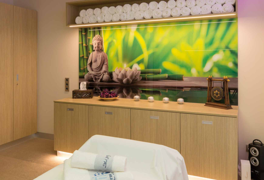 Ein hell erleuchteter Therapieraum mit Cremefarbenden Teppich. Im unteren Bereich des Bildes befindet sich eine Massageliege bedeckt mit einem weißen Laken und weißen Handtüchern mit der Aufschrift SaarowTherme. Im Hintergrund befindet sich eine helle Holzkommode mit Dekorativen Elementen wie einer Taschentücherbox, eine Schale mit Lila Blumen, 4 Teelichterständer und einem Chinesischen Gong Becken. Über der Kommode hängt ein hell grünes Bild mit einem Buddah, darüber ein Hängeschrank mit weißen Handtüchern