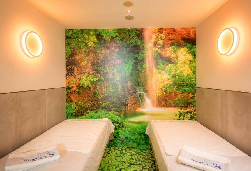 Ein Wellnessraum mit zwei Packungsliegen rechts und links im Bild. Beide Liegen sind mit weißem Bettlaken und ein SaarowTherme Handtuch drapiert. Im Hintergrund und auf den Fußboden des Wellnessraumes ist ein Vollflächiges Bild von einem grünen Wasserfall zu sehen. An den Wänden im Raum befinden sich links und rechts eine Runde Wandlampe die im Kreis nach Innen und nach Außen die Wand beleuchtet.