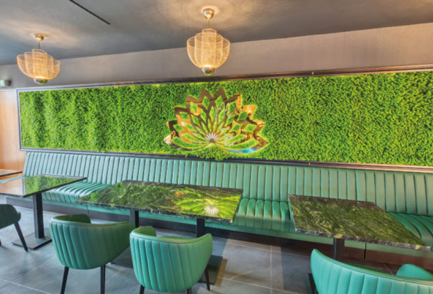Ein kleiner Ausschnitt von dem Baderestaurant Lotus Gourmet in der SaarowTherme. Im Vordergrund befinden sich grüne Leder bezogene Sessel an eckigen Tischen mit einer Platte aus schwarz weiß marmoriertem Marmorplatte. Der Hintergrund wird von einer langen Sitzbank und einem Wandbild aus grünem Moos mit dem Lotus Logo des Restaurants verzeirt.