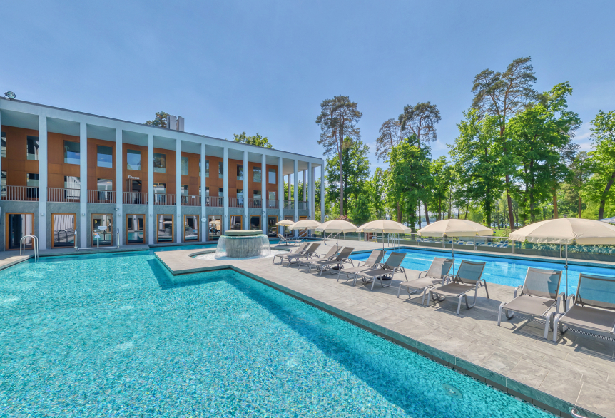 Außenbereich des Thermalsolebeckens  SaarowTherme am Tag mit plätschernden Strömungskanal, türkisfarbenen Natursolebecken, graue Sonnenliegen mit weißen aufgespannten Sonnenschirmen. Im Hintergrund ein Teil des Hauptgebäudes mit hellbrauner Gebäudefront und weißen Säulen im Vordergrund. Strahlend blauer Himmel. 