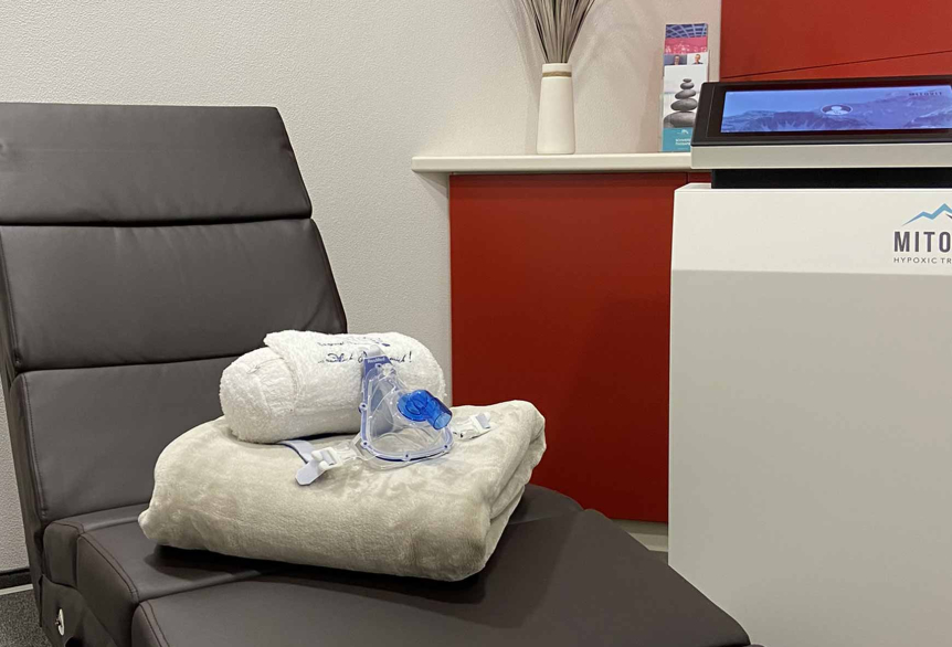 Gepolsterte Liege aus dunklem braunen Leder. Darauf liegt ein weißes Handtuch, weiße Nackenrolle und eine Atemmaske für die Sauerstofftherapie. Im Hintergrund ist eine rote Schranktür mit einem Flyerständer mit Werbung der SaarowTherme zu sehen. Im Vordergrund ist die Motivit Maschine für die Sauerstofftherapie zu sehen.