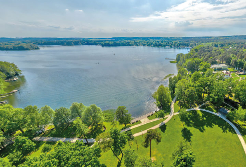 Luftaufnahme vom Scharmützelsee in Bad Saarow. Blauer Himmel, Sonnenschein und das funkeln auf dem Wasser der Scharmützelsees. Am Ufer sieht man einen Ausschnitt vom grünnen Kurpark Bad Saarow mit seinen Uferwanderwegen.