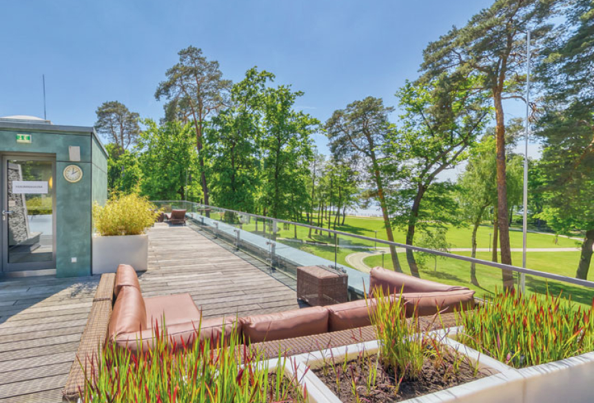 Großzügige Dachterrasse mit bequemen Lounge-Sitzgruppen und dekorativen Pflanzkästen. Die Terrasse bietet einen weiten Blick auf grüne Bäume und den nahegelegenen Scharmützelsee. Links befindet sich ein Zugang mit Glasfassade.