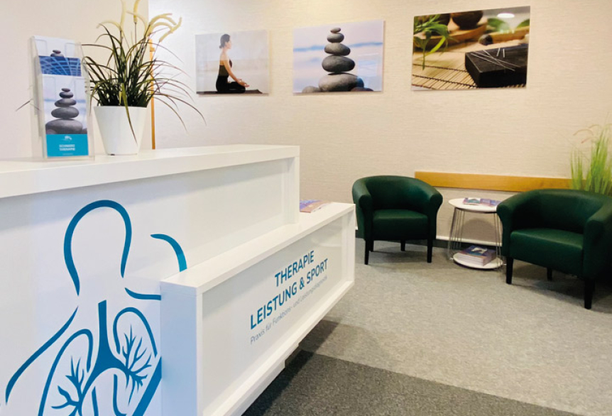 Empfangsbereich einer Arztpraxis. Im Vordergrund steht ein großer weißer Tresen mit dem Schriftzug Therapie, Leistung und Sport – Arztpraxis der SaarowTherme. Der Fußboden ist mit einem hellgrauen Teppich belegt, die Wände mit einer hellen Strukturtapete versehen und an der Wand hängen große Bilder mit alternative Therapien abgebildet, wie Akupunktur. Zwei Sesseln stehen an der Rückwand und dazwischen einen kleinen Beistelltisch mit Broschüren.