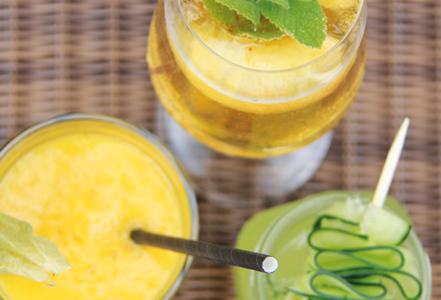 Drei farbenfrohe Sommergetränke in Gläsern – ein gelber Smoothie mit Physalis, ein goldener Drink mit Minze und Ananas sowie ein grüner Cocktail mit Gurkenspirale – auf einem geflochtenen Untergrund.