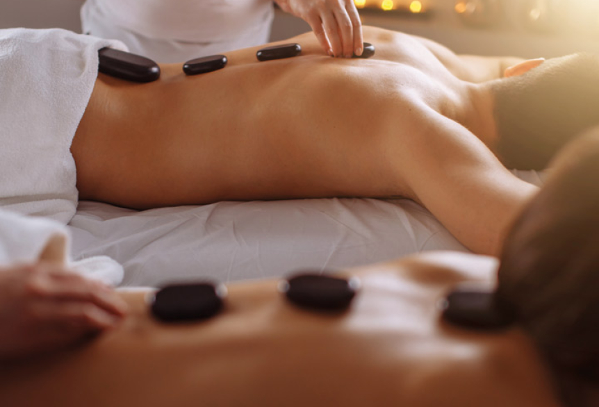 Mann und Frau liegen jeweils auf eine Massageliege. In Bauchlage mit den Händen über Ihren Köpfen entspannen Sie, währen Therapeuten auf den Rücken der beiden warme Massagesteine legen. Im Hintergrund leuchten Kerzen, die für eine entspannte Stimmung sorgen.
