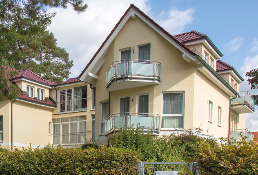 Zweistöckiges hellgelbes Wohnhaus mit roten Dächern und kleinen Balkonen, umgeben von Grünpflanzen und Bäumen unter blauem Himmel.