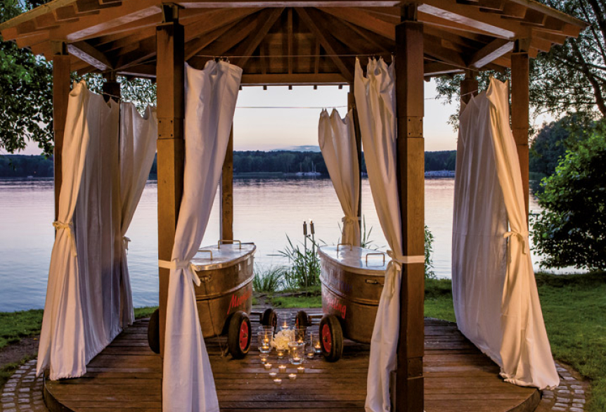 Pavillon am See mit seitlichen Vorhängen drapiert für Privatsphäre. In der Mitte vom Pavillon stehen zwei Metallwannen auf Räder, die mit einem Deckel verschlossen sind. Zwischen den Wannen sind mehrere Kerzen auf den Boden verteilt worden, die für romantische Stimmung sorgen. 
