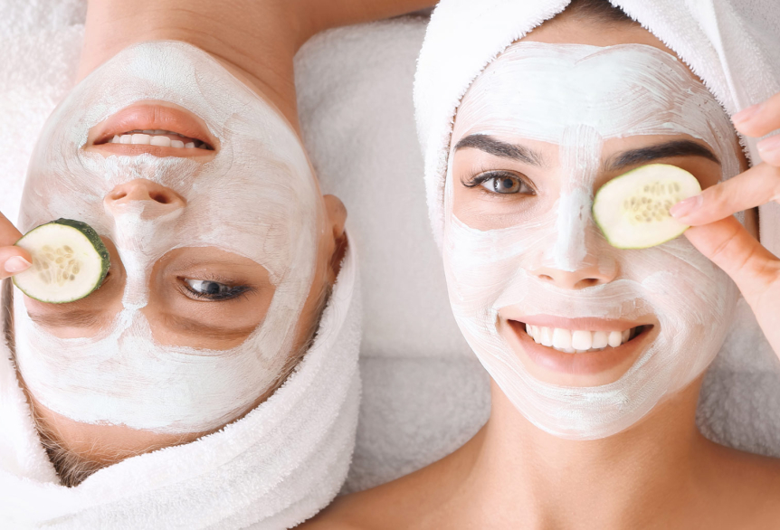 Zwei junge Frauen liegen auf dem Rücken auf einem Bett. Haare sind eingehüllt in einem Handtuch und auf dem Gesicht tragen Sie eine Gesichtsmaske und halten sich jeweils eine frische Gurke vor dem Auge.