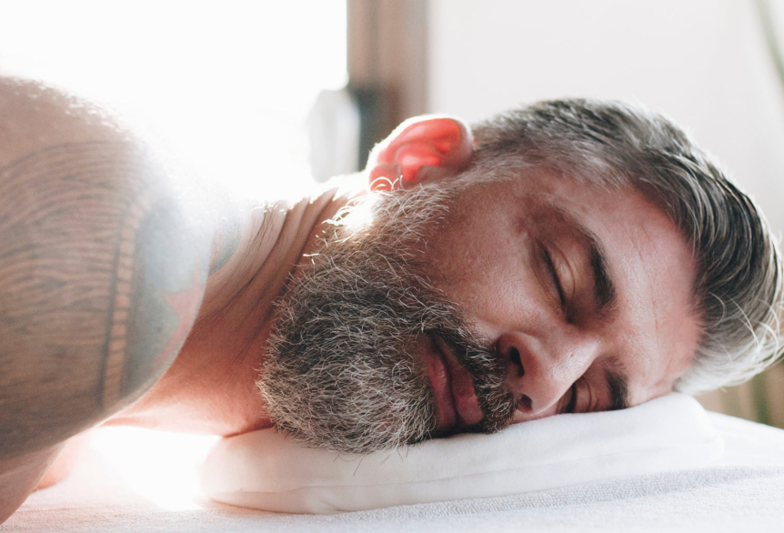 Mann mit Vollbart liegt in Bauchlage auf einer Massageliegen. Augen zu genießt er eine Wellnessbehandlung. 