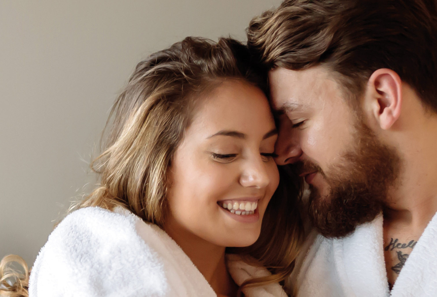 Junges Pärchen liegt auf einer Wellnesslounge mit kuscheligen Kissen in einem Beigeton. Das Paar umarmt sich und lächelt sich gegenseitig an. Sie tragen beide einen weißen Bademantel.