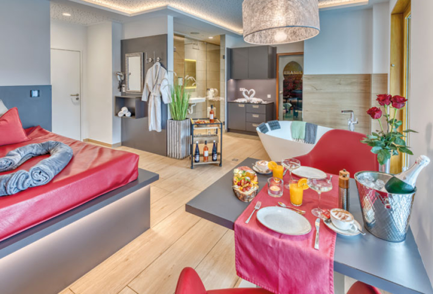 Modernes Wellness-Zimmer mit rotem Doppelbett, romantisch gefalteten Handtüchern, Esstisch mit gedecktem Frühstück, Champagner und Rosen. Im Hintergrund sind eine freistehende Badewanne und ein offenes Bad mit Bademänteln sichtbar.